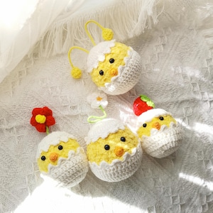 Może przedstawiać: Cztery szydełkowe kurczaki w skorupkach jajek, każdy z innym wzorem. Jeden kurczak ma kwiat na górze, drugi truskawkę, a trzeci żółtą antenę. Kurczaki są wszystkie białe z żółtymi dziobami i łapkami.