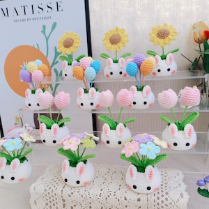 Puede incluir: Una colección de macetas de conejito de crochet con varias decoraciones florales y de globos. Las macetas son blancas con orejas rosas y se exhiben en un estante. Las flores son rosas, moradas y azules, y los globos son de colores pastel.