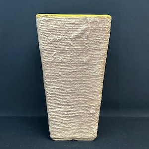 May include: A tall, tapered vase with a textured, beige, stone-like exterior surface. The vase features a distinct, flat, bright yellow rim along the top edge, contrasting with the neutral, rough-hewn finish of the main body of the vessel.