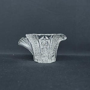 May include: A clear glass creamer featuring a faceted star pattern on the body, a fan-shaped handle on one side, and a small pouring spout on the opposite side. The glass has a textured surface with geometric detailing throughout the exterior.