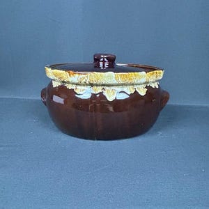 May include: A round, dark brown ceramic pot with a matching lid and two small side handles. The upper rim of the pot and the edge of the lid feature a decorative, textured drip glaze in a creamy, light tan color that contrasts with the smooth body.