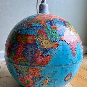 May include: A vintage globe with a colorful map of the world, featuring continents and countries in various hues. A white light fixture is attached to the top of the globe. The globe sits on a wooden floor.