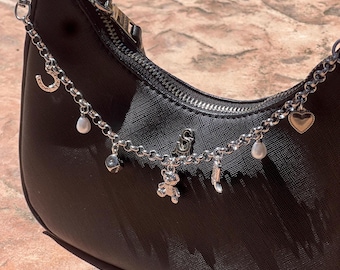 May include: Black handbag with a silver chain strap adorned with various charms. Charms include a horseshoe, a heart, a teddy bear, a pearl, and a small cube. The bag has a zipper closure and a silver chain.
