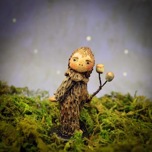 May include: A small, brown, textured figurine of a creature with a round face, rosy cheeks, and dark eyes. The figure has a shaggy, bark-like body and holds a thin branch with two small, round, light-colored objects resembling marshmallows.