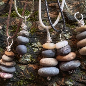 May include: Six necklaces featuring pendants made of stacked, smooth, multi-colored river stones. Each pendant is secured with silver-toned wire wrapping and hangs from a simple cord in various colors including black, brown, white, and olive green.
