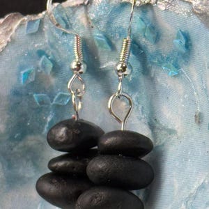 May include: Earrings featuring stacked black stones, suspended from silver-tone hooks. Each earring showcases a series of smooth, dark pebbles, creating a natural, minimalist aesthetic. The earrings have decorative silver accents at the base.