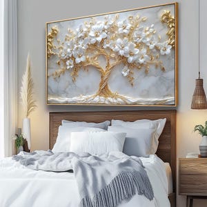 May include: A framed wall art piece featuring a 3D relief of a tree with white flowers and gold accents. The artwork is set against a marble-like background and framed in gold. The piece is displayed above a bed with white bedding.