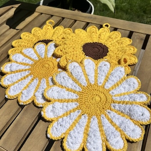 May include: Three round, crocheted flower-shaped pot holders. Two feature white petals with a yellow center, while one features yellow petals with a dark brown center. Each piece includes a small loop at the top for hanging, showcasing detailed yarn work.