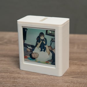 May include: A white rectangular photo frame with a slot on top, displaying a family photo. The photo shows a man and a child. The frame is sitting on a wooden surface.