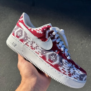 May include: A low-top sneaker featuring a red, white, and grey geometric pattern across the upper. The shoe includes a white leather Nike swoosh, white laces, and a white rubber sole with the text AIR embossed on the side heel area.