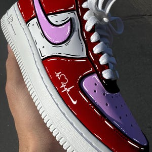 May include: Customized red, white, and pink Air Force 1 sneakers. The shoe features a heart and heartbeat design on the side, with a pink Nike swoosh. White laces and a white sole complete the design.