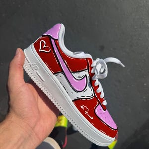 May include: Red and pink Air Force 1 sneakers with a white sole. The sneakers feature a heart design and a pink Nike swoosh. The laces are white.