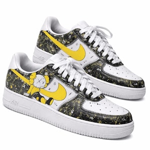 May include: A pair of white sneakers featuring a black and yellow paint splatter pattern, bright yellow swoosh logos, and a cartoon character graphic with X-shaped eyes on the side panels. The shoes have white laces and white rubber soles.
