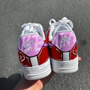 May include: Pair of red and white Nike Air Force 1 sneakers with custom pink and white accents. The heels feature the Nike Air logo and a heart design. White laces and soles complete the look.