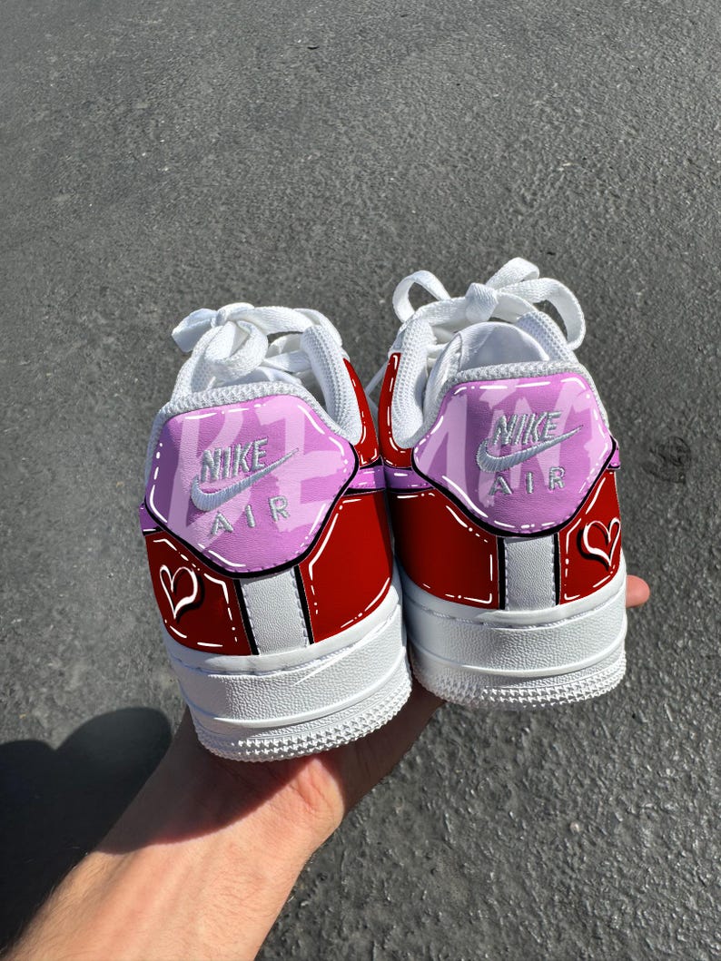 May include: Pair of red and white Nike Air Force 1 sneakers with custom pink and white accents. The heels feature the Nike Air logo and a heart design. White laces and soles complete the look.