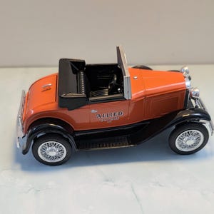 May include: A die-cast model of an orange and black vintage convertible car with wire-spoke wheels. The side of the vehicle features the text ALLIED VAN LINES in black lettering. The interior is black with a steering wheel and a folded top.