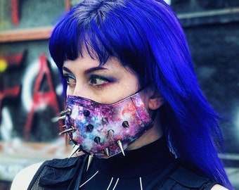 May include: A person wearing a black tactical vest with multiple pockets and a black mesh panel. They are also wearing a black mask with a galaxy print and silver spikes. The person has blue hair and is standing in front of a graffiti wall.
