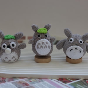 May include: Three handmade crochet Totoro figures in grey, white, and green. Each figure has large eyes and a white belly. The figures are displayed on a white, curved platform, showcasing the craftsmanship of the plush toys.
