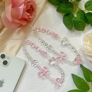 May include: Two clear beaded phone charms with pink bow accents. The charms are strung with clear beads and feature small pink bow-shaped beads. A white smartphone with a silver Apple logo is visible.