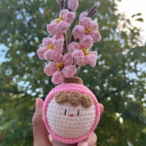 May include: A crocheted vase with a pink and white face and brown hair, holding a pink flower bouquet. The vase is on a wooden base.