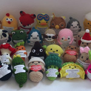 May include: A selection of colourful, crocheted amigurumi figures, including animals, food items, and whimsical characters. Each figure has a small tag with text. The figures are in various colours, such as brown, yellow, pink, and green.