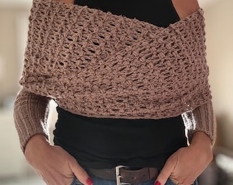 May include: A light brown, open-weave, crocheted shoulder wrap featuring a textured, lattice-like pattern. The garment has long, ribbed sleeves and is worn over a black sleeveless top, paired with blue denim jeans and a brown leather belt.