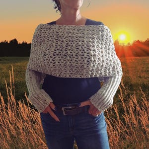 May include: A light grey, speckled, off-the-shoulder crochet shrug featuring a textured, open-stitch pattern across the chest and shoulders, with long, ribbed sleeves that extend down to the wrists, worn over a dark blue shirt and blue jeans.
