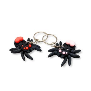 Könnte beinhalten: Zwei Spinnen-Schlüsselanhänger aus schwarzem Material. Eine Spinne hat leuchtend rote Augen und ein rotes Herz auf dem Rücken, während die andere zartrosa Augen und ein passendes rosa Herz hat. Jede Spinne ist an einem runden Metallring befestigt.