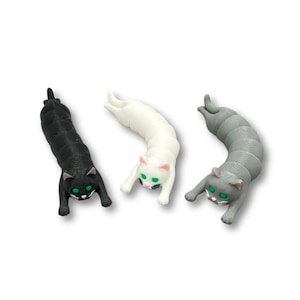 Puede incluir: Tres figuras con forma de gato en negro, blanco y gris. Cada gato tiene ojos verdes y detalles rosas en las orejas y la nariz. Los gatos están en posición de gateo, con el cuerpo arqueado.
