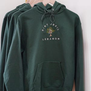 Op de afbeelding: Een donkergroen hoodie met een wit geborduurd ontwerp met een boom en de tekst "Bint Jbeil Libanon".