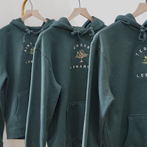 Puede incluir: Tres sudaderas con capucha verde oscuro con diseños bordados y la palabra "Líbano". Cada sudadera cuelga de una percha de madera. Las sudaderas tienen un bolsillo delantero y una capucha con cordón. El texto incluye nombres de ciudades libanesas.