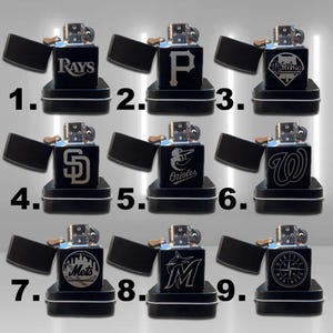 May include: A collection of nine black lighters, each with a different team logo engraved in silver. The lighters are displayed open, revealing their internal mechanisms. Each lighter sits on a small, black, rectangular box.