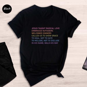 May include: Black t-shirt with the text "JESUS TAUGHT RADICAL LOVE EMBRACED OUTSIDERS WELCOMED SINNERS CALLED US TO SHOW GRACE TO HEAL, NOT TO HATE TO INCLUDE, NOT TO EXCLUDE IN HIS NAME, WALK HIS WAY" in a rainbow gradient. Black trousers and a smartphone are also visible.