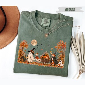 May include: A moss green short-sleeve t-shirt featuring a graphic print of a rabbit, a ghost, a goose, and a fox dressed in witch hats and capes. The scene includes autumn trees, sunflowers, a full moon, and bats. The shirt has a Comfort Colors label.