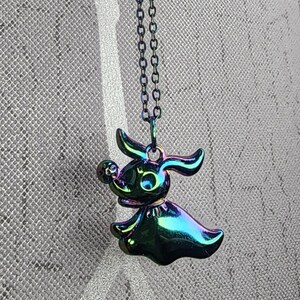 Puede incluir: Un collar negro iridiscente con un dije de Zero, el perro fantasma de Pesadilla antes de Navidad. El dije está en una pose de vuelo con una cabeza grande y redonda y un cuerpo largo y delgado.