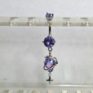 Puede incluir: Un piercing para el ombligo de color plateado con tres acentos de piedras preciosas moradas. La gema superior es pequeña y redonda. La gema del medio es un corte grande y redondo. La gema inferior es un diseño en forma de planeta con un colgante en forma de estrella.