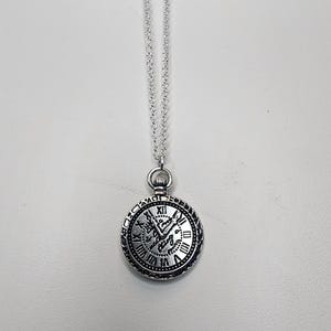 Puede incluir: Collar con colgante de reloj de bolsillo plateado. El colgante redondo presenta números romanos y detalles ornamentados alrededor de la esfera. El collar tiene una cadena delicada y un cierre en la parte superior.