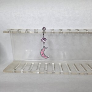 May include: A silver-toned belly button ring featuring a small pink gem at the top, a larger round pink gem in the center, and a dangling crescent moon charm. The moon charm is accented with pink enamel and small white star-shaped cutouts.