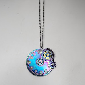 Może przedstawiać: Naszyjnik ze srebrnym łańcuszkiem i wisiorem w kształcie tęczowej, opalizującej tarczy zegara z trzema mniejszymi kołami zębatymi. Tarcza zegara ma czarne rzymskie cyfry.