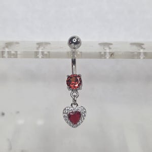 May include: A silver belly button ring with a large, round, red gemstone and a dangling heart-shaped charm. The heart is red with a border of small, clear stones. The top ball is also silver.