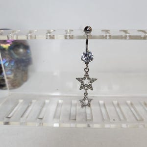 May include: Silver belly button ring with a dangling star design. The top features a clear, round gem, with two star-shaped charms hanging below. The stars are embellished with small, clear stones.