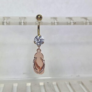 May include: A rose gold belly button ring with a clear, round gemstone and a dangling flip-flop charm. The flip-flop charm is detailed with small, clear gemstones. The top ball is gold.