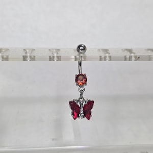 May include: A silver belly button ring with a dangling butterfly charm. The charm features a red oval-shaped gem and a red butterfly with gem accents. The top gem is also red and oval-shaped.