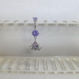 Könnte beinhalten: Ein silberfarbener Bauchnabelpiercing mit einem kleinen violetten Edelstein oben, einem größeren runden violetten Edelstein in der Mitte und einem hängenden Lotusblüten-Anhänger unten, der einen tropfenförmigen violetten und rosafarbenen Stein enthält.