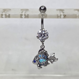 May include: A silver belly button ring featuring a fish charm. The fish is detailed with blue and white gems, and the ring has a large, clear, round gem at the top. The fish charm adds a decorative element.