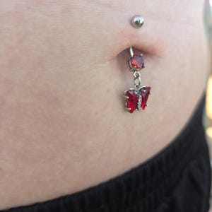 May include: A silver-toned navel piercing ring featuring a round red gemstone at the top and a dangling butterfly-shaped charm set with red stones. The jewelry is inserted into the navel, with a smooth metal ball securing the top of the barbell.