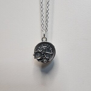 Op de afbeelding: Een zilveren ketting met een ronde hanger met een klok met Romeinse cijfers en tandwielen. De hanger is bevestigd aan een zilveren ketting.