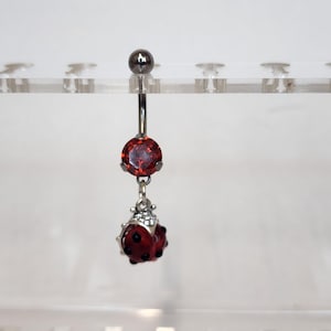 May include: A silver belly button ring with a large, round, red gemstone and a dangling ladybug charm. The ladybug charm is red with black spots and small silver accents. The top of the ring has a small silver ball.