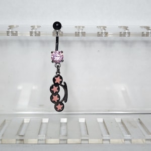 Può includere: Anello per ombelico nero con una gemma rosa e un ciondolo a forma di stella. Il ciondolo presenta tre stelle rosa su sfondo nero. La sfera superiore è nera.