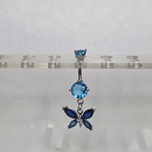 May include: A silver belly button ring featuring a butterfly charm. The ring has a light blue gem at the top, a larger light blue gem in the center, and a butterfly charm with dark blue and white gems.